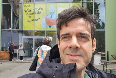 Na imagem, Maoel Candeias homem de cabelos curtos e escuros e cavanhaque está parado em frente a um prédio com uma placa que diz “TEATRO AN WERK”. Ele está vestindo uma jaqueta azul sobre uma camisa xadrez vermelha e tem uma expressão neutra no rosto. O teatro parece ser um espaço moderno, possivelmente renovado, com grandes janelas que permitem a entrada de luz natural no interior. Não há outras pessoas visíveis nas imediações do homem.